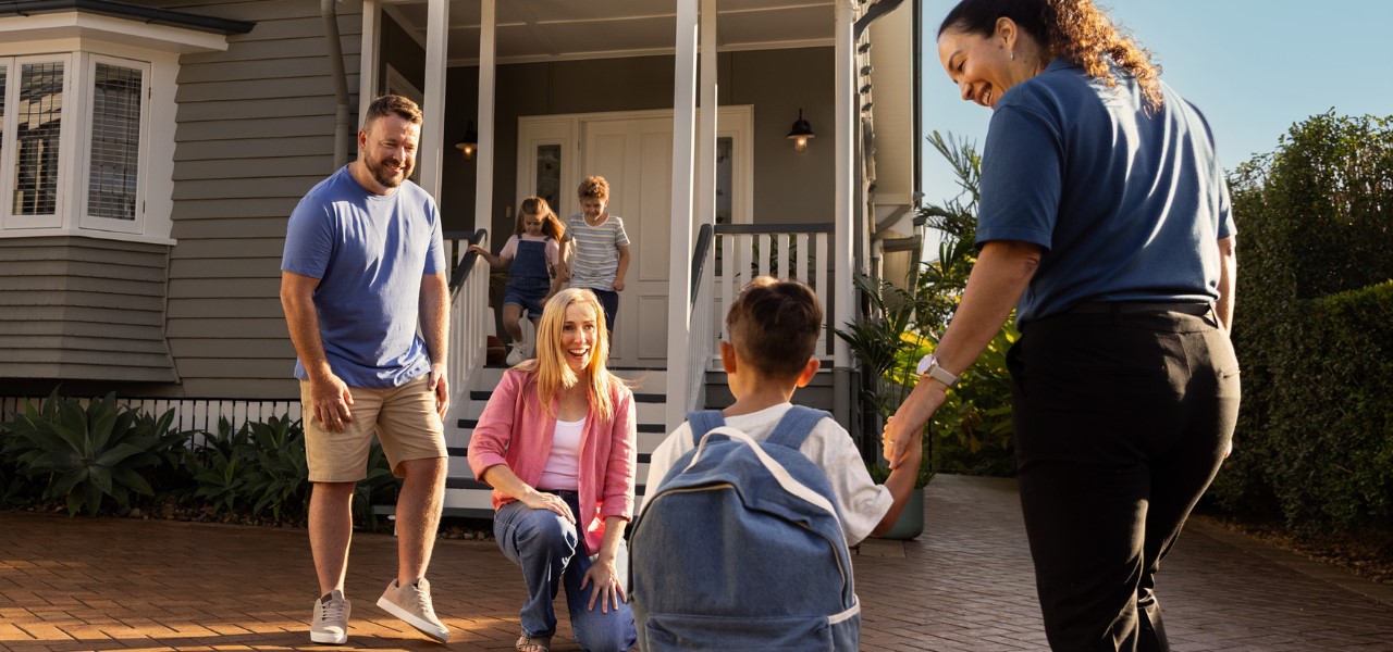 A couple and 2 children stand in front of a house. In front of them is a small child with a backpack holding the hand of a middle aged female.