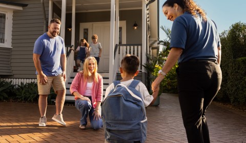 A couple and 2 children stand in front of a house. In front of them is a small child with a backpack holding the hand of a middle aged female
