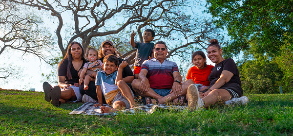 A family of 8 First nations people sit together on a picnic blanket on a grassy lawn beneath a large tree with spreading branches. The family group includes several adults and children, with 1 child standing behind the seated group. Green trees and a bright sky form the background in an outdoor park setting.