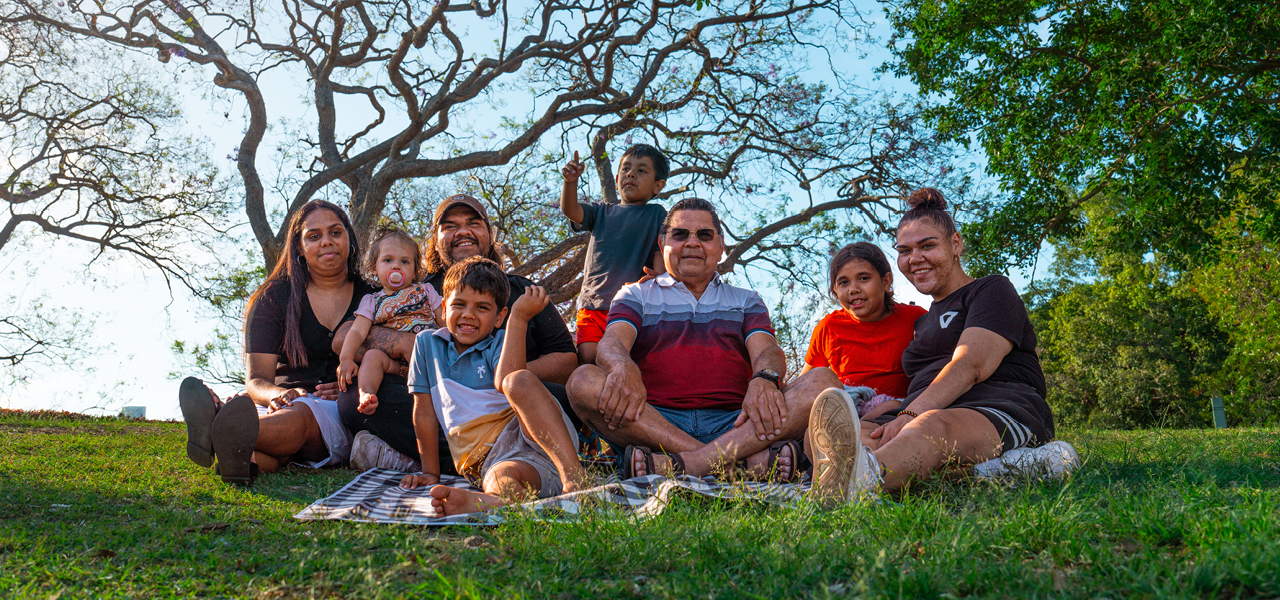 A family of 8 First nations people sit together on a picnic blanket on a grassy lawn beneath a large tree with spreading branches. The family group includes several adults and children, with 1 child standing behind the seated group. Green trees and a bright sky form the background in an outdoor park setting.